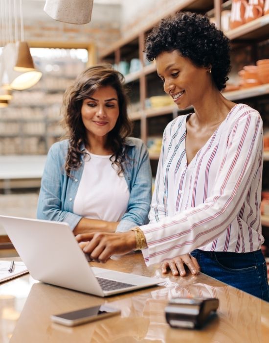 Two women in a retail setting collaborate using a laptop, with a payment terminal and smartphone on the counter.