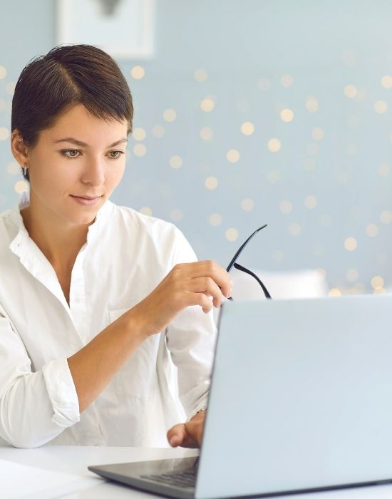 A young professional woman sitting at a desk holding glasses and working on a laptop.